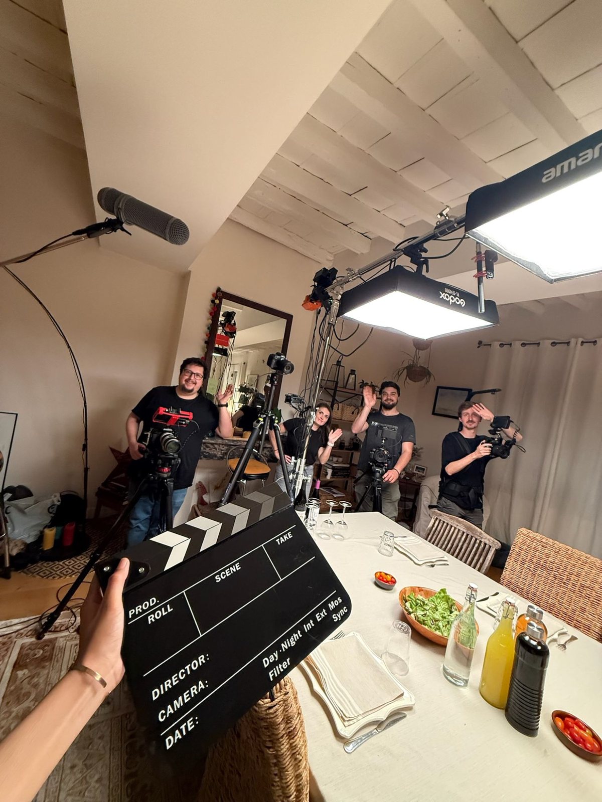 Un tournage en appartement : l’équipe salue derrière le clap, caméras et lumières en place.